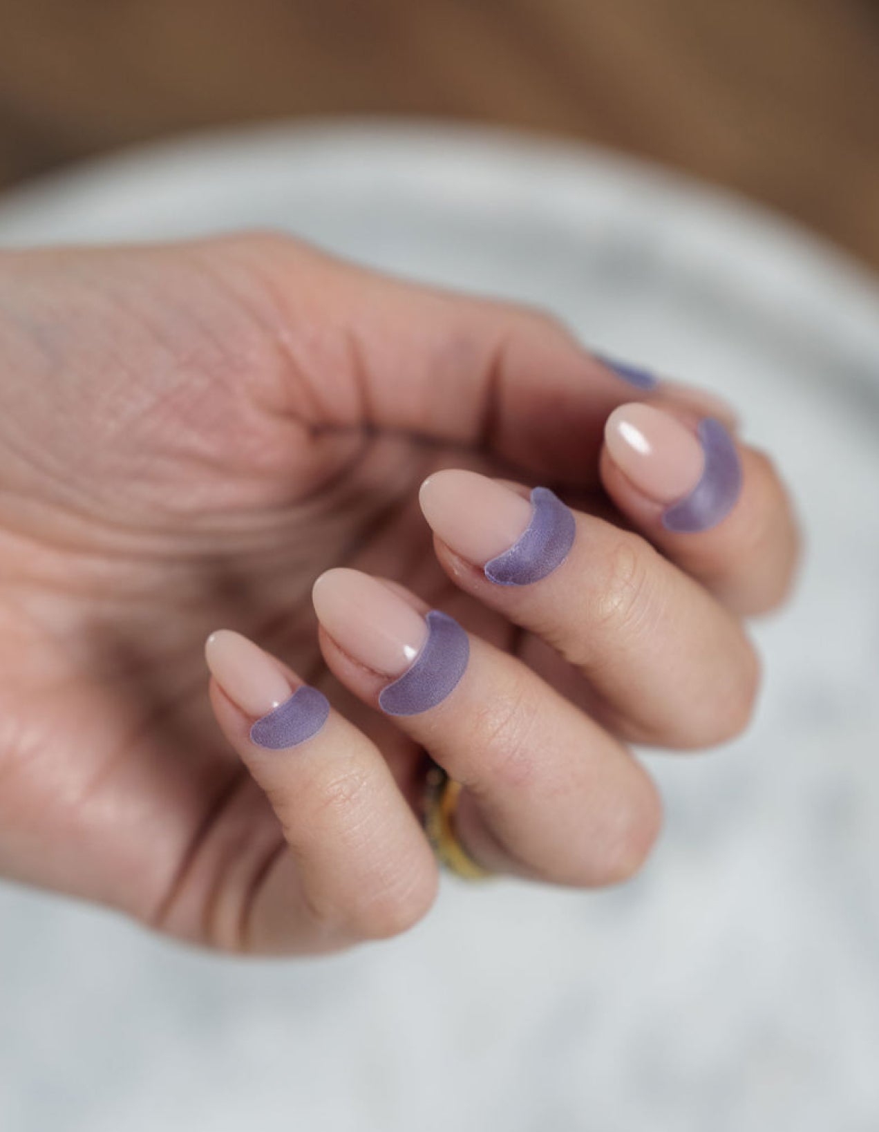 Close-up of a hand wearing French Lilac Cutipatch cuticle patches with neutral nail polish on a blurred background.